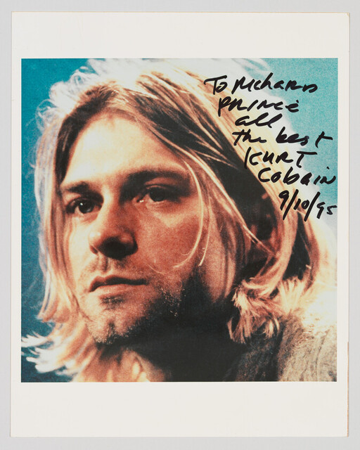 Close-up portrait of a man with shoulder-length blond hair and a black handwritten note.