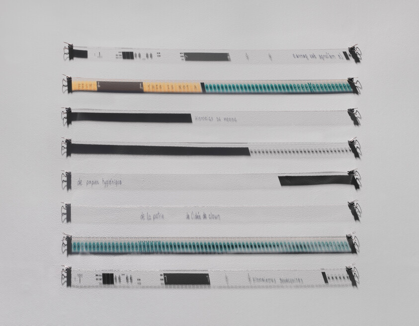 Seven long labeled strips with clips arranged horizontally on a white wall, some with colored patterns.