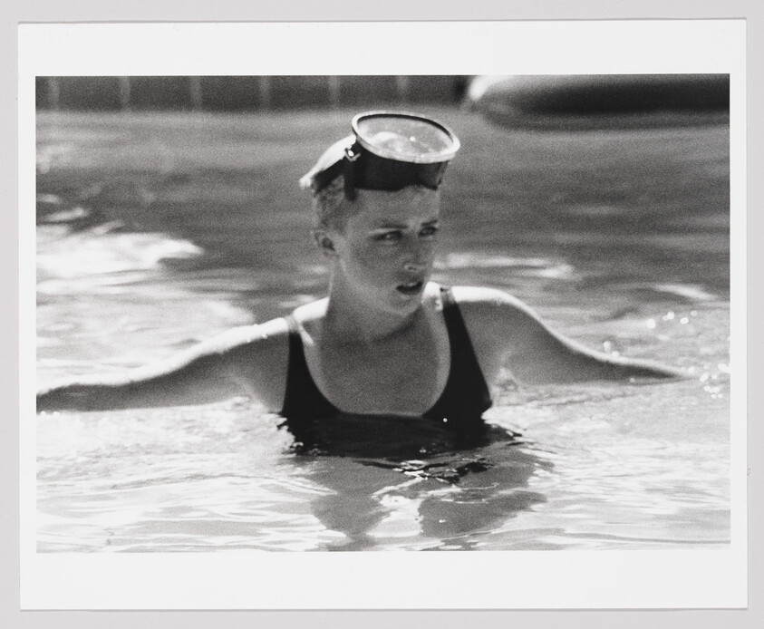 Young woman wearing swim goggles rests in a pool with arms outstretched.