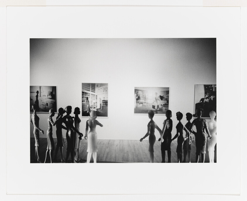 A line of mannequins in a gallery facing framed photographs on a white wall.