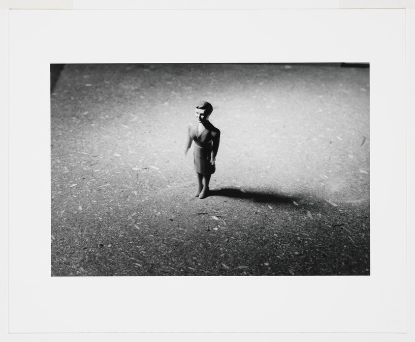 A small plastic doll stands alone on rough ground, casting a long shadow to the right.