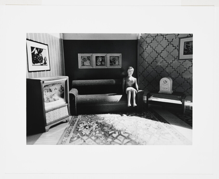 A small mannequin sits on a couch inside a vintage dollhouse living room with patterned rug.
