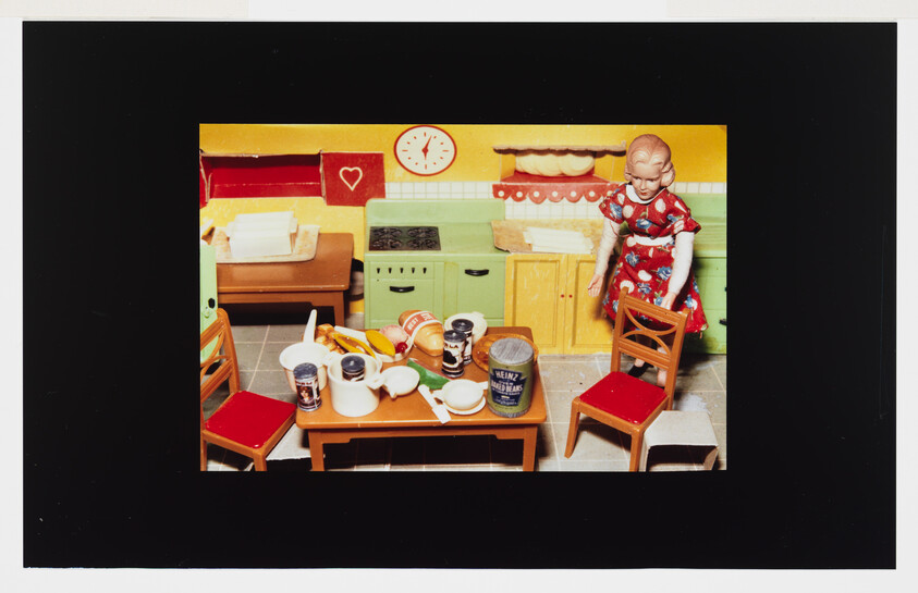 A doll in a red dress stands beside a small kitchen table cluttered with dishes and food.