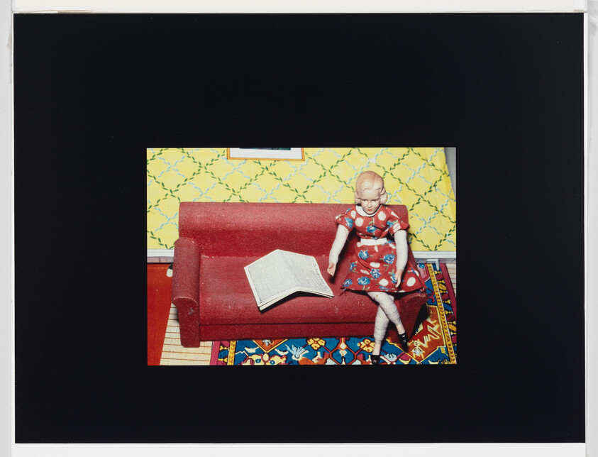A vintage doll in a red dress sits on a red couch beside an open book.
