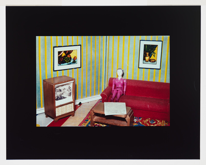 A small plastic doll sits on a red sofa in a vintage miniature living room with a coffee table.