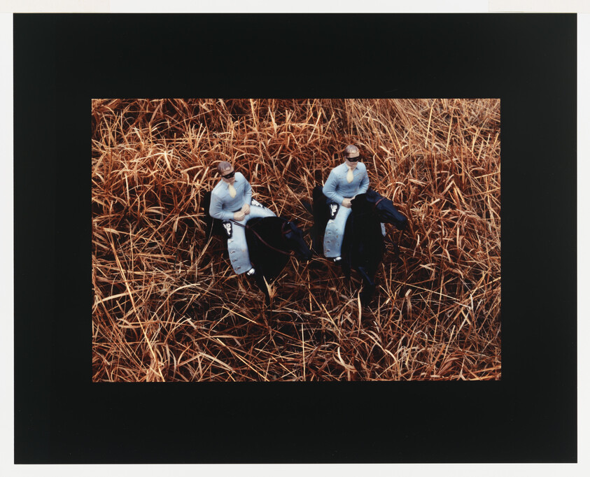 Two riders wearing light shirts sit on dark horses in tall dry grass.