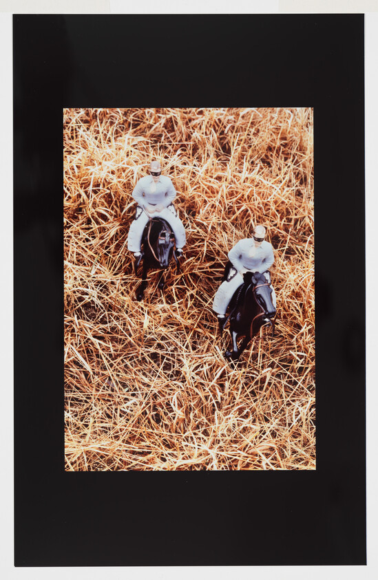 Two toy horseback riders in white outfits ride through tall dry brown grass.