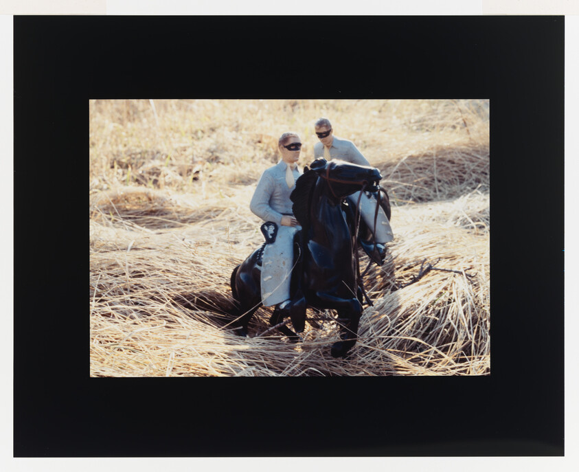 Two masked riders guide a black horse through tall dry grass while riding side by side.