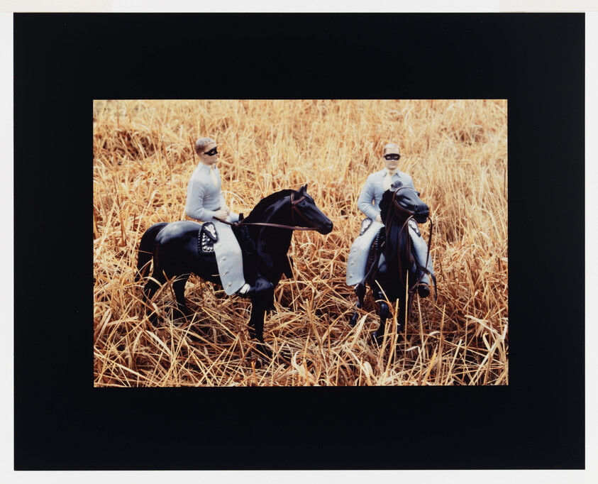Two toy riders in white outfits sit on black horses standing in dry, tall grass.