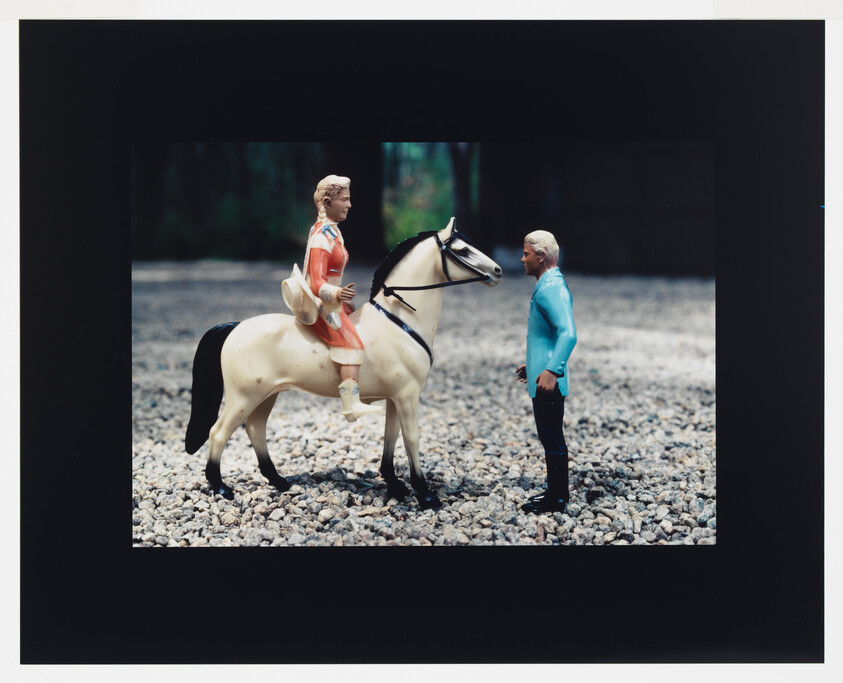 A toy rider on a white horse faces a standing toy figure on gravel.