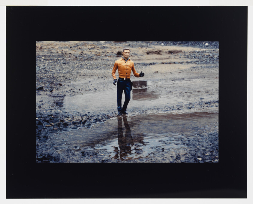 A toy action figure in an orange shirt walks through a muddy puddle with its reflection visible.