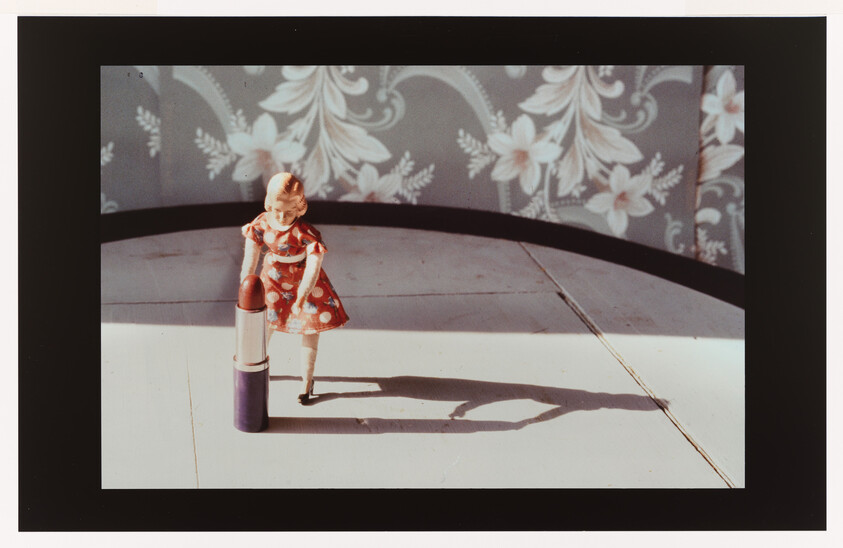 A small doll in a red dress stands beside an oversized lipstick, casting a long shadow.
