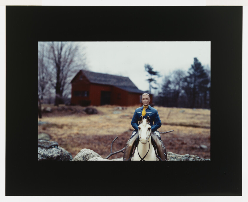 Toy cavalry officer on a white horse sits in front of a red barn in a field.