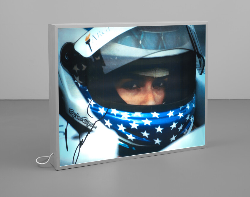 A framed photograph of a person wearing a racing helmet and a bandana with an American flag pattern is displayed on a stand. The helmet features a visor and sponsorship branding, and there appears to be an autograph on the photo.