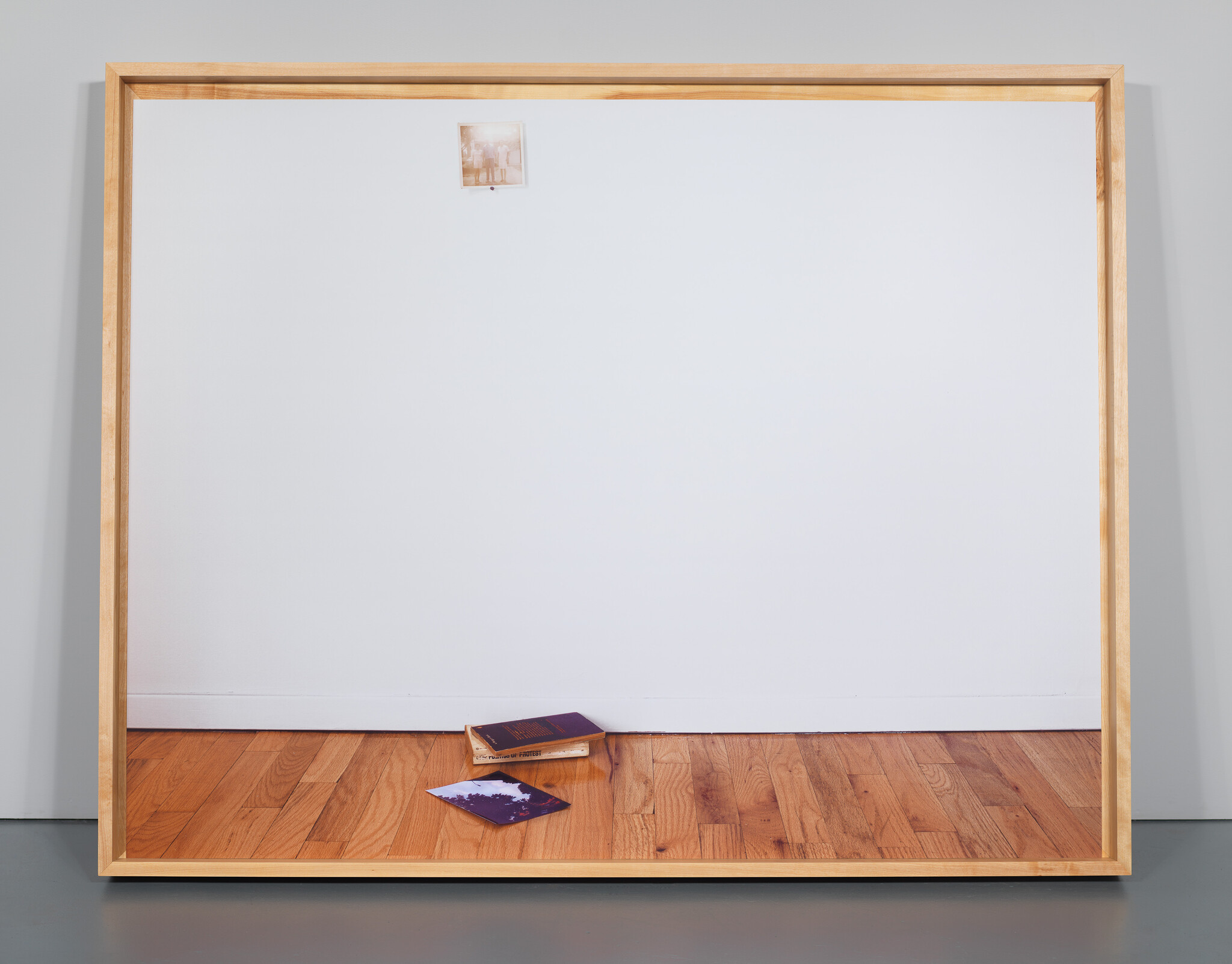 A large framed white wall with a small pinned photo and two books lying on the wooden floor.