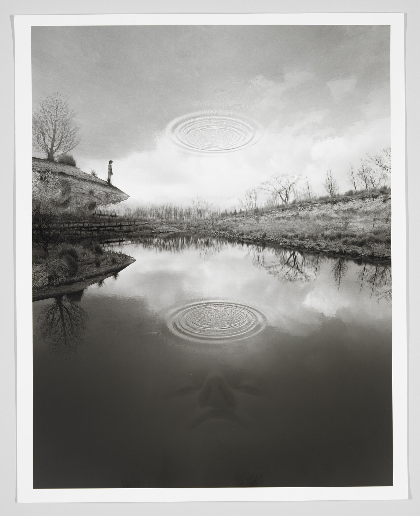 A lone person stands on a riverbank looking at concentric ripples and a faint face reflected in the water.
