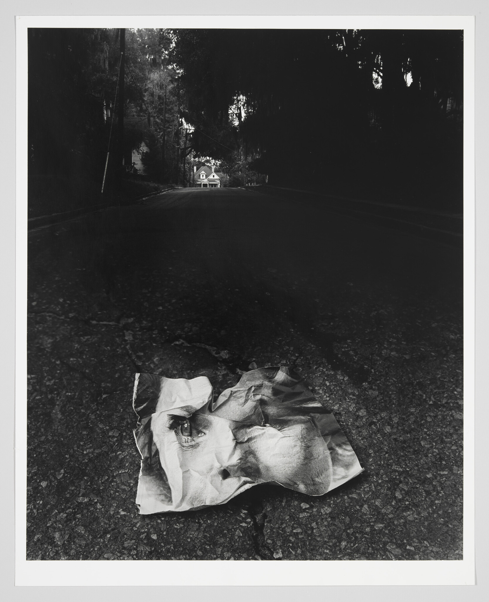 A crumpled paper with a printed face lies on an empty asphalt road toward a distant house.
