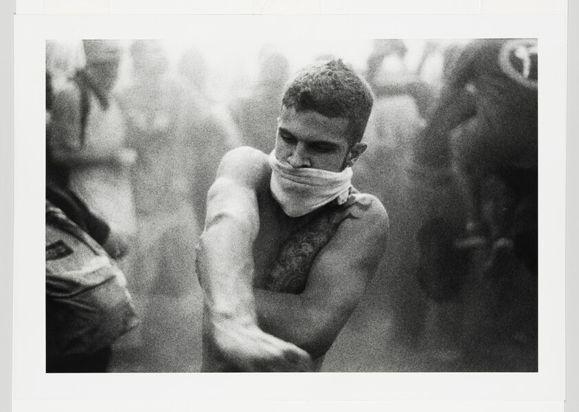 Shirtless young man with cloth over his mouth pulls his arm amid a smoky crowd.