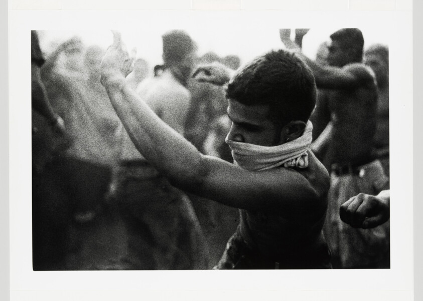 Young shirtless man with cloth covering his mouth raises his arms amid a crowded, smoky gathering.