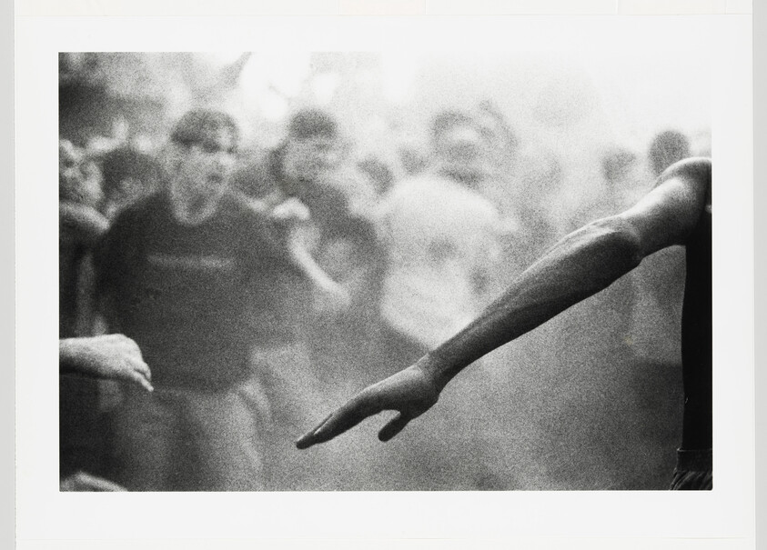 A person extends an outstretched arm toward another hand amid a crowded, smoky scene.
