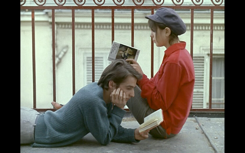 A young man lies reading while a young woman sits behind him reading on a balcony.