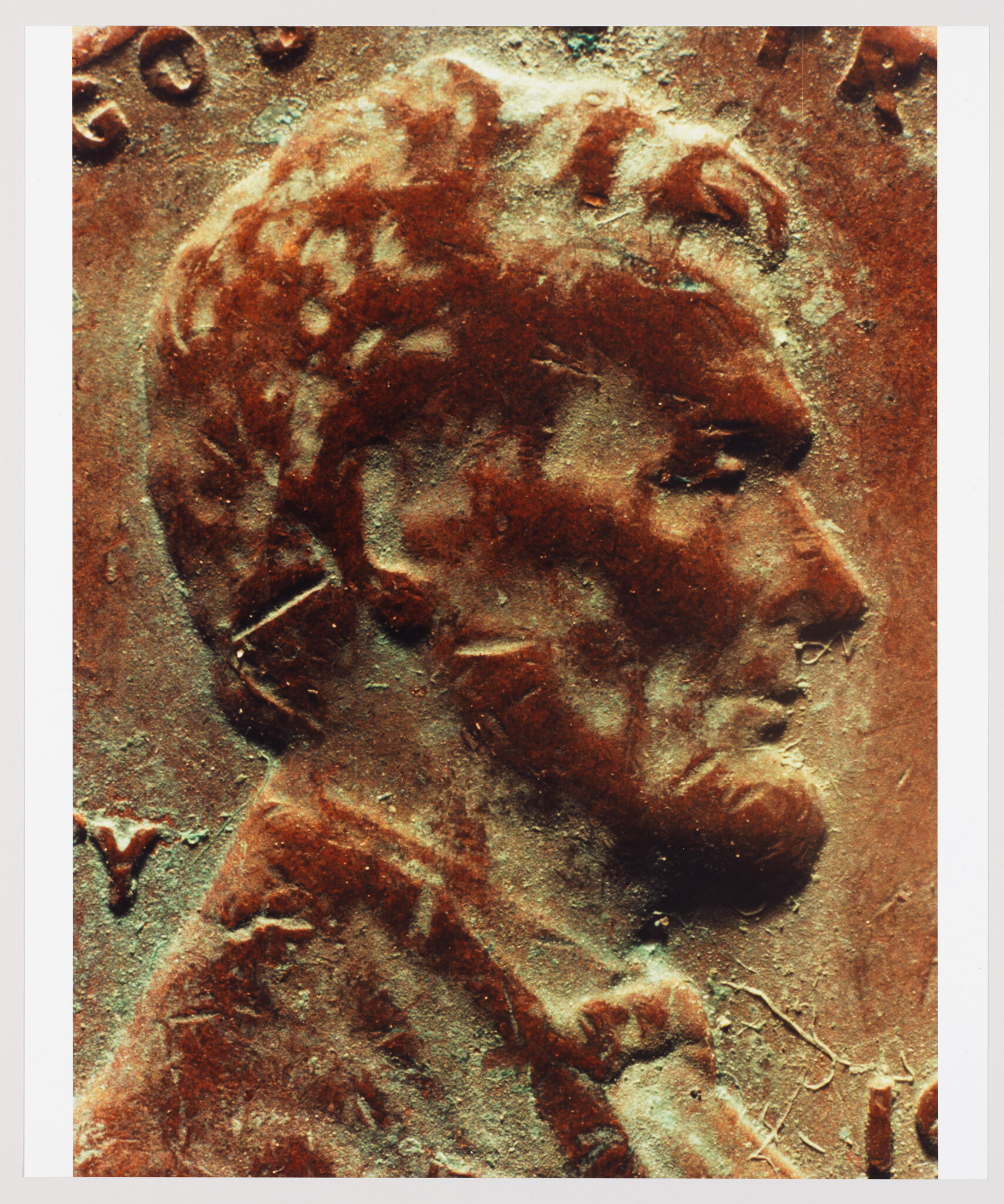 Close-up of a weathered copper coin featuring the profile of a male figure with short hair, possibly a historical figure or leader, with inscriptions and symbols partially visible around the edges. The coin's surface shows signs of oxidation and wear.