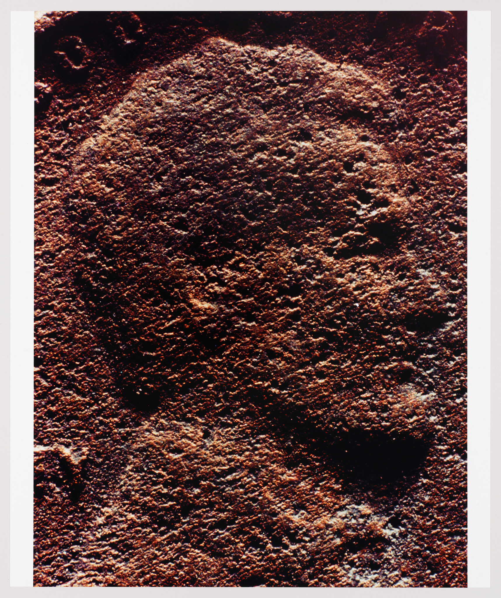 Close-up photo of a rocky surface with a rough texture, showcasing various shades of brown and red, possibly indicative of iron-rich minerals or Martian soil. The lighting creates a high contrast between the bright and shadowed areas, emphasizing the uneven terrain.