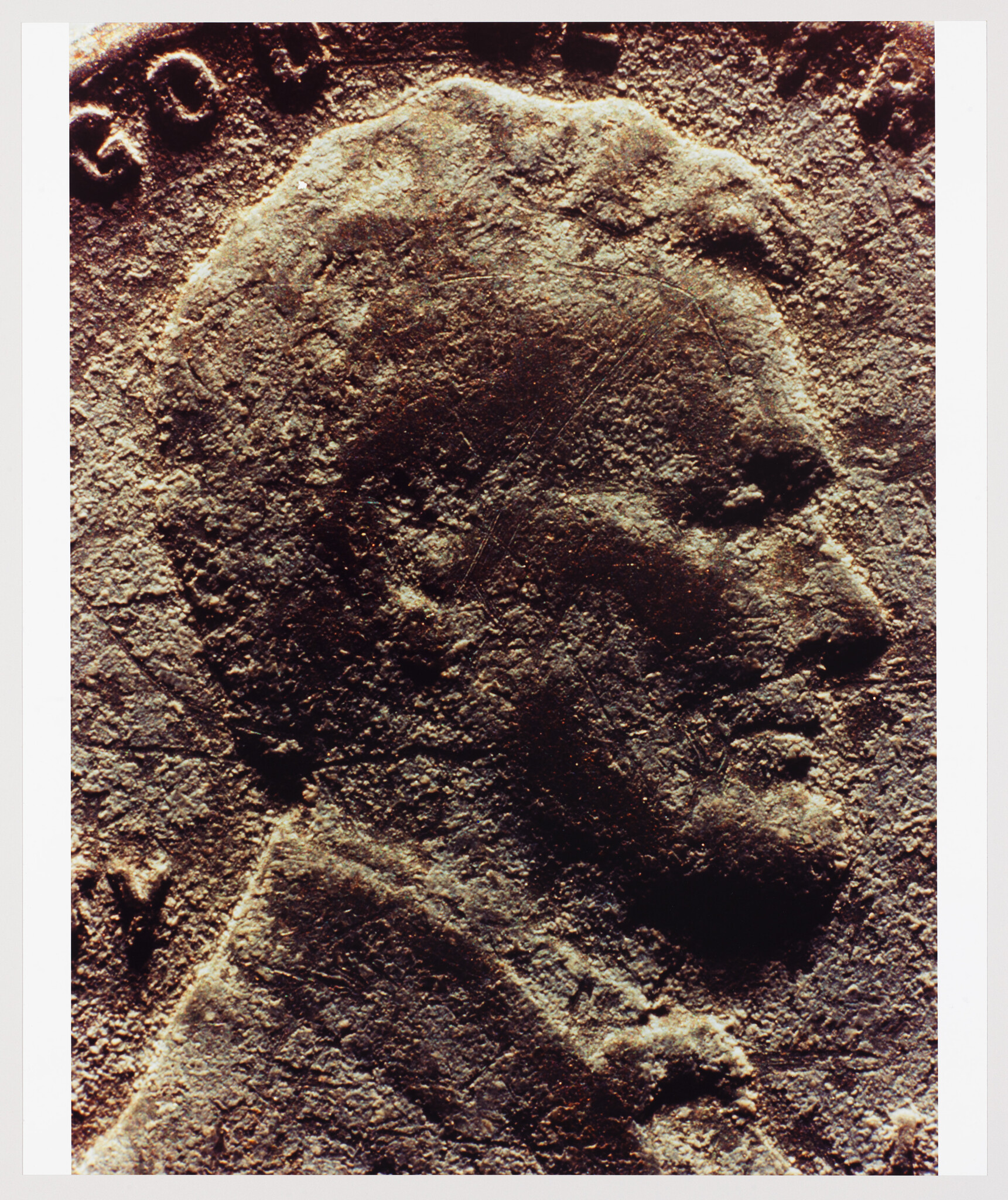 A close-up of a weathered bas-relief carving depicting a male profile with classical features, possibly from an ancient coin or monument, with visible inscriptions partially cropped at the top edge.