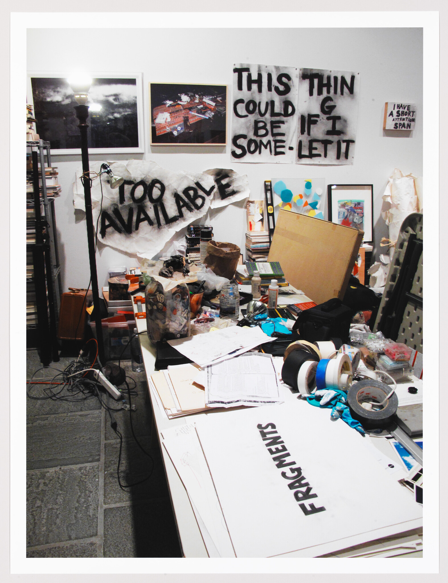 Cluttered artist workspace with scattered papers, tape, and signs reading "Too Available" and "Fragments."