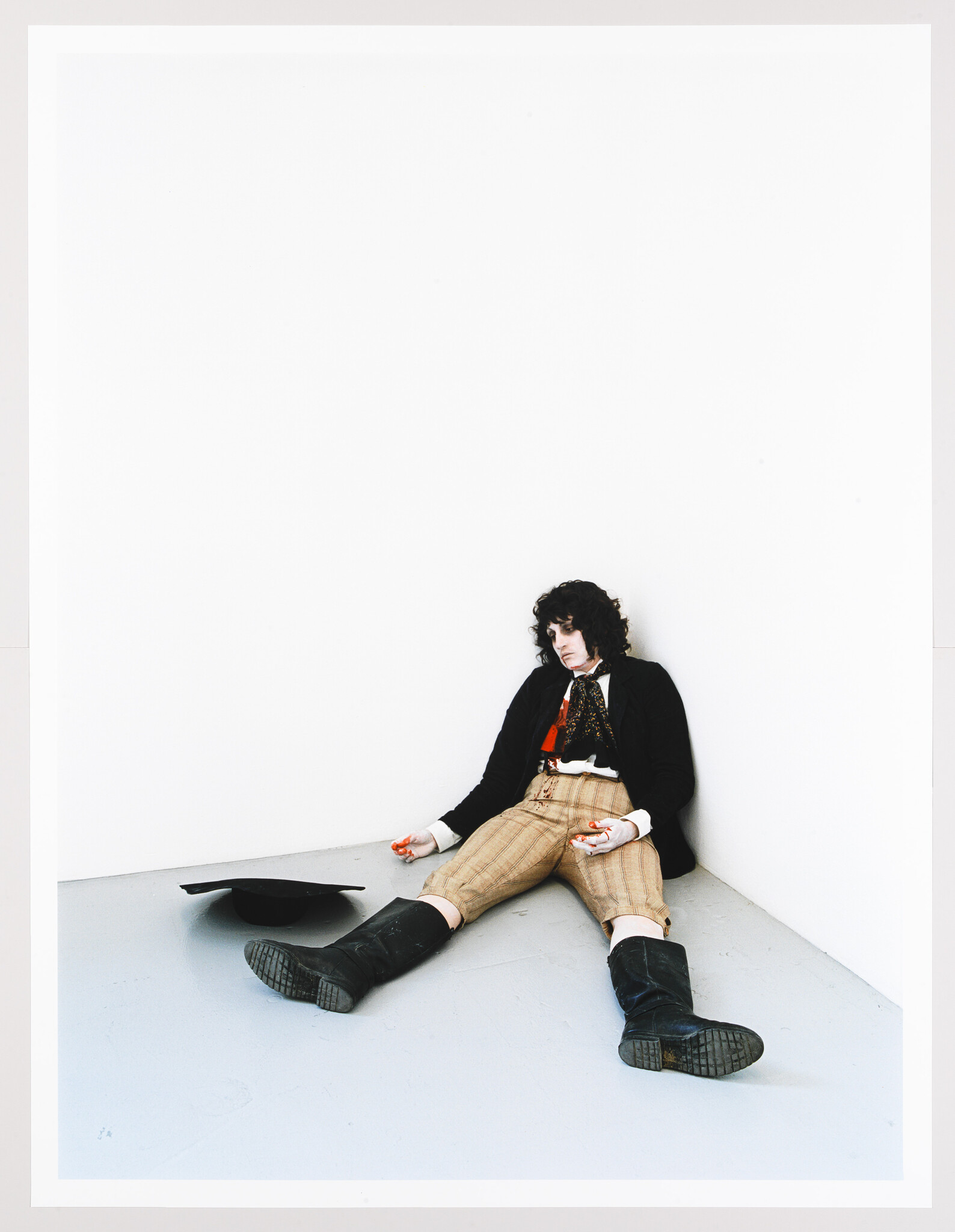 A young person in vintage clothing slumps against a white corner, hat fallen beside them.