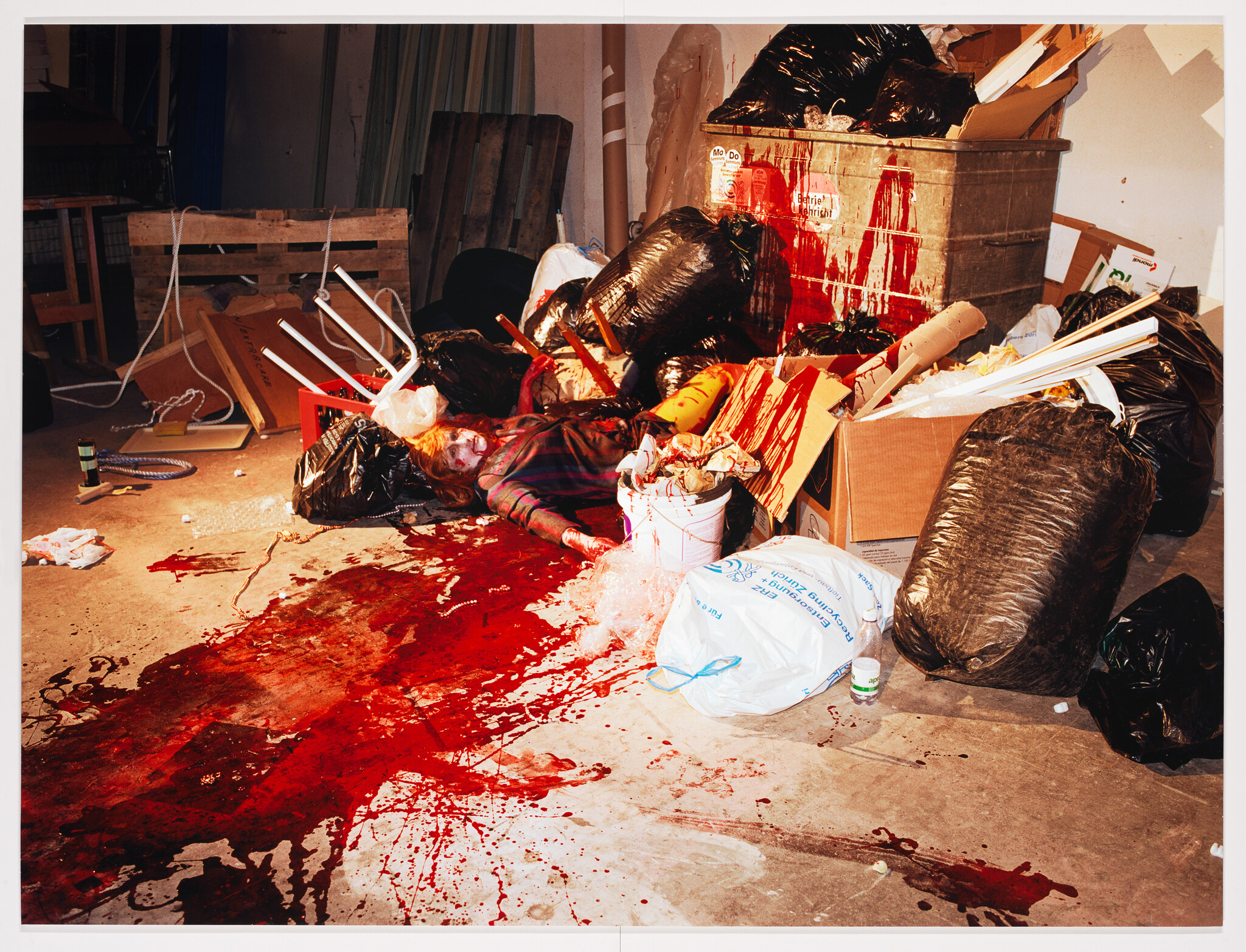 A person lies motionless among trash with a large pool of blood spreading across the floor.