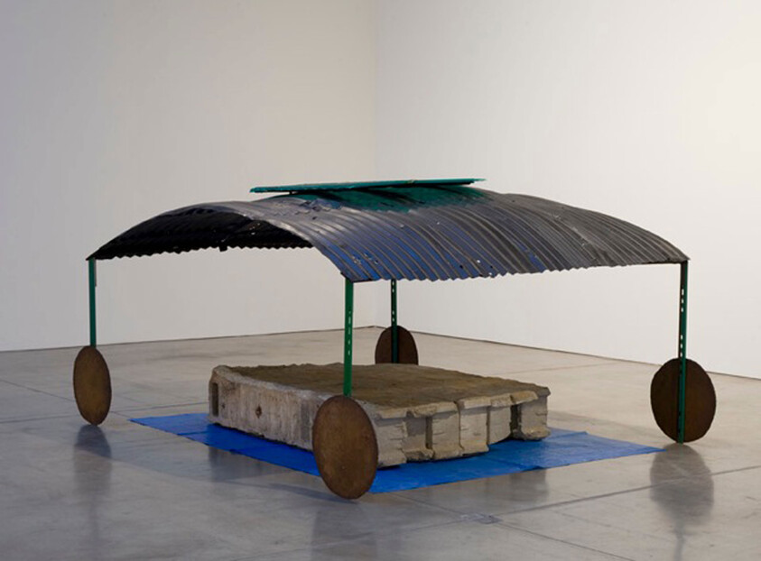 Corrugated metal canopy with green posts and round wheels sheltering a concrete slab on a blue tarp.