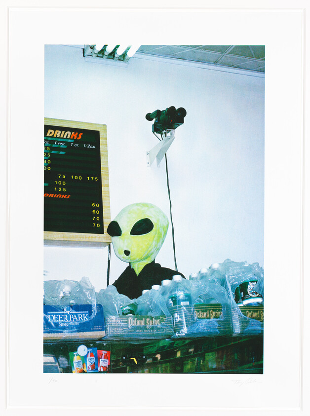 A person wearing an oversized alien mask with large black eyes is standing behind a stack of bottled water at a store. Above them is a menu board listing drink prices, and to the right, a security camera is mounted on the wall. The image has a candid, snapshot quality and includes the artist's signature and edition number at the bottom.
