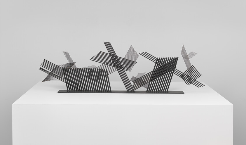 An abstract metal sculpture of intersecting black slatted panels displayed on a white pedestal.