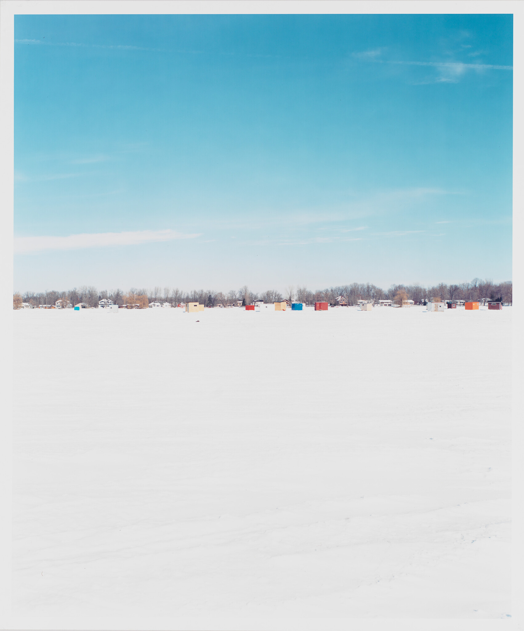 A row of colorful ice fishing huts sits on a wide frozen lake under a pale blue sky.