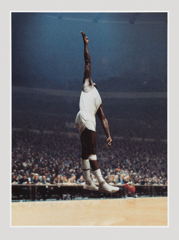 Basketball player in mid-air, reaching high with one arm extended, wearing a white uniform, with a large crowd in the background.