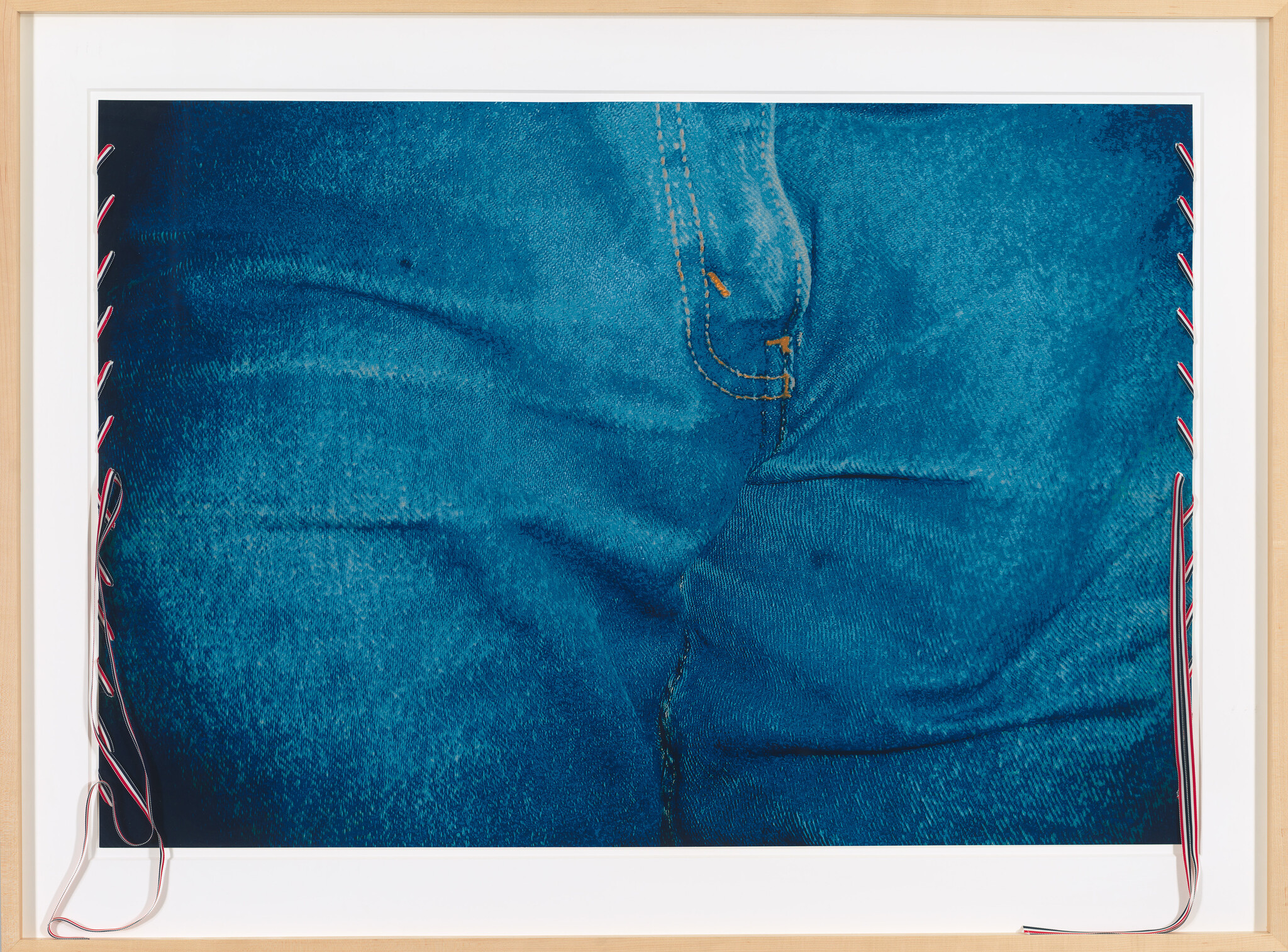 A close-up photograph of a section of blue denim fabric with visible texture and seams, framed in a light wooden frame with red and white cords laced through the left side, creating an artistic presentation.