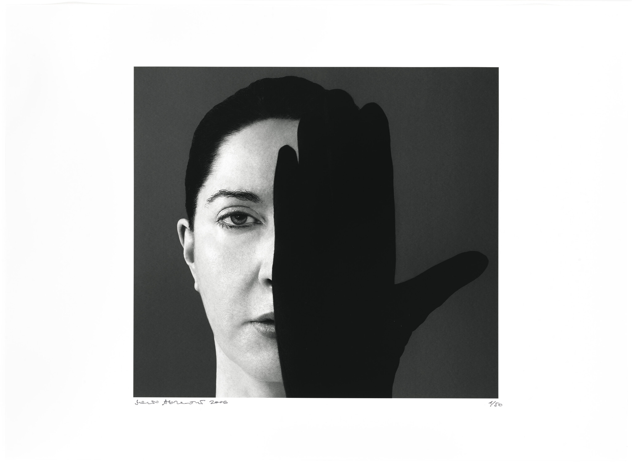 Black and white photograph of a woman's face partially obscured by a silhouette of a hand in the foreground. The image has a stark contrast between the light and dark areas, with the woman's expression appearing neutral. There is text at the bottom left of the image, likely indicating the artist's signature and the year, and a fraction at the bottom right, possibly denoting the print number of the artwork.