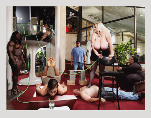 A crowded living room shows a blonde woman standing on a chair holding a green hose while others lie and watch.