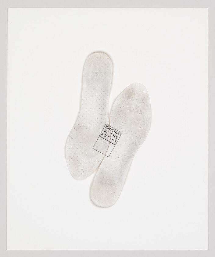 Two worn white shoe insoles lie flat, one stamped with the words "Touched by the artist".