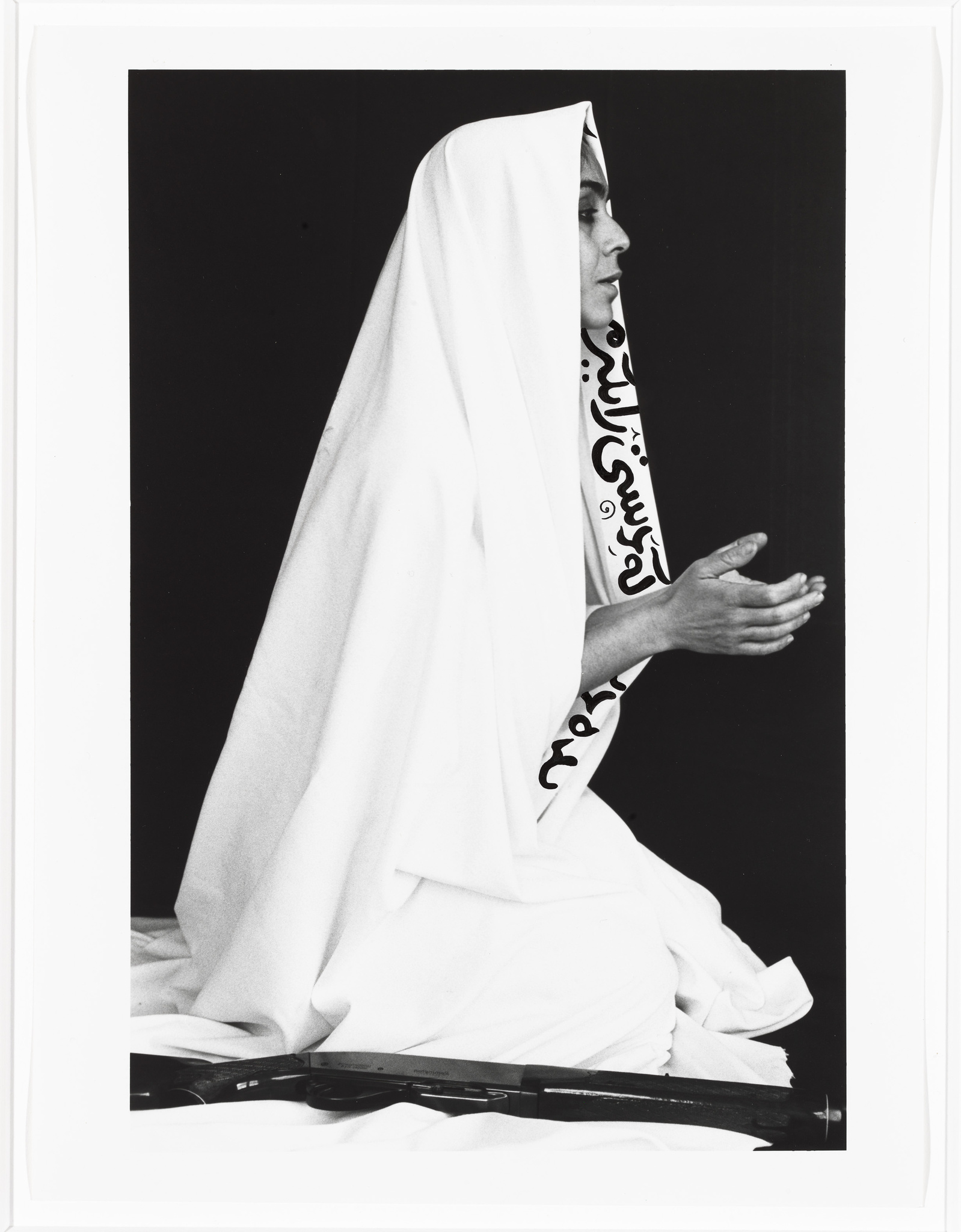 Seen from a profile view of a Iranian woman who is facing the right is wearing a white veil with Farsi text, has her hands in a prayer position, and has a gun near her bended knees 