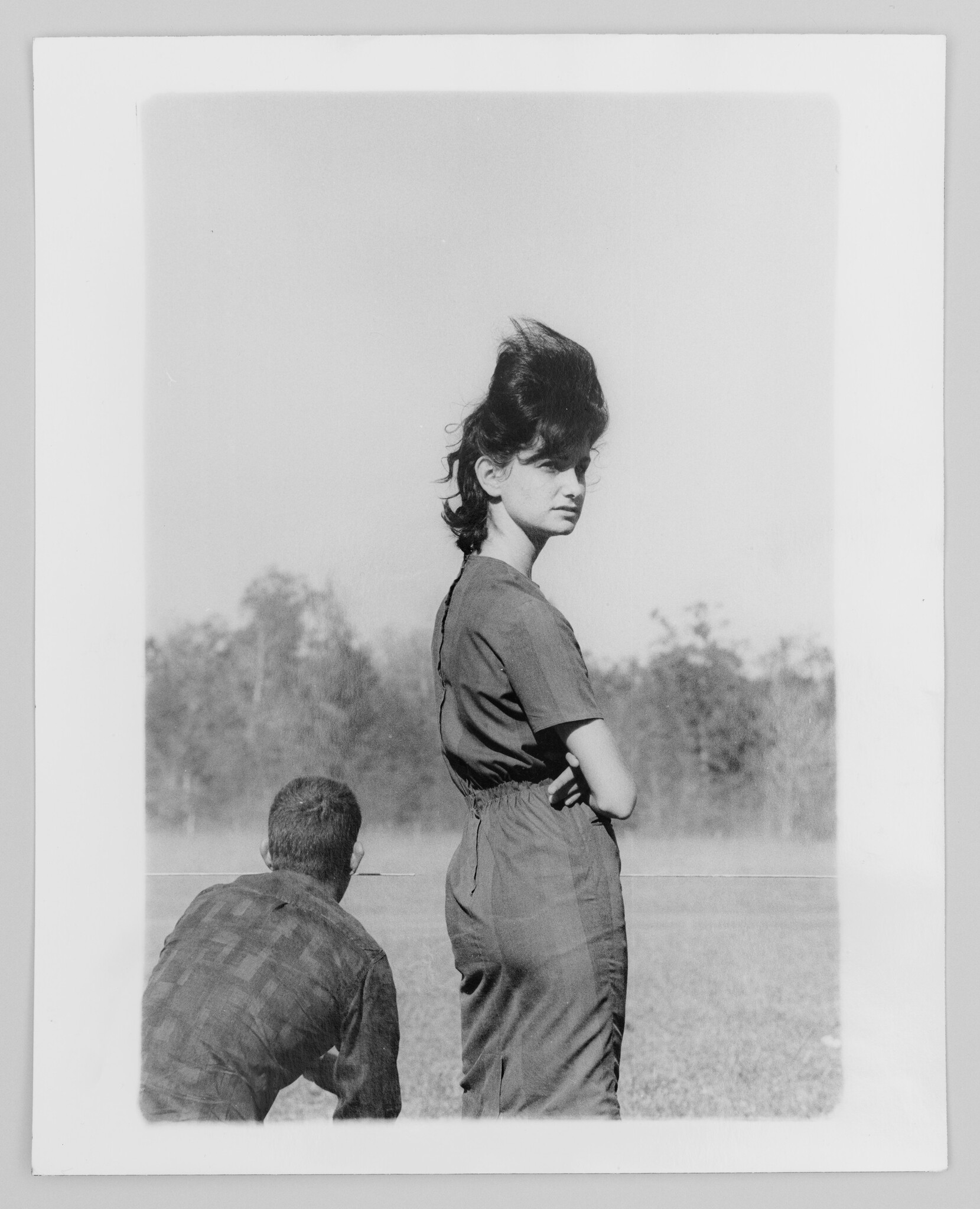 Young woman stands with arms crossed looking over a field while man crouches nearby.