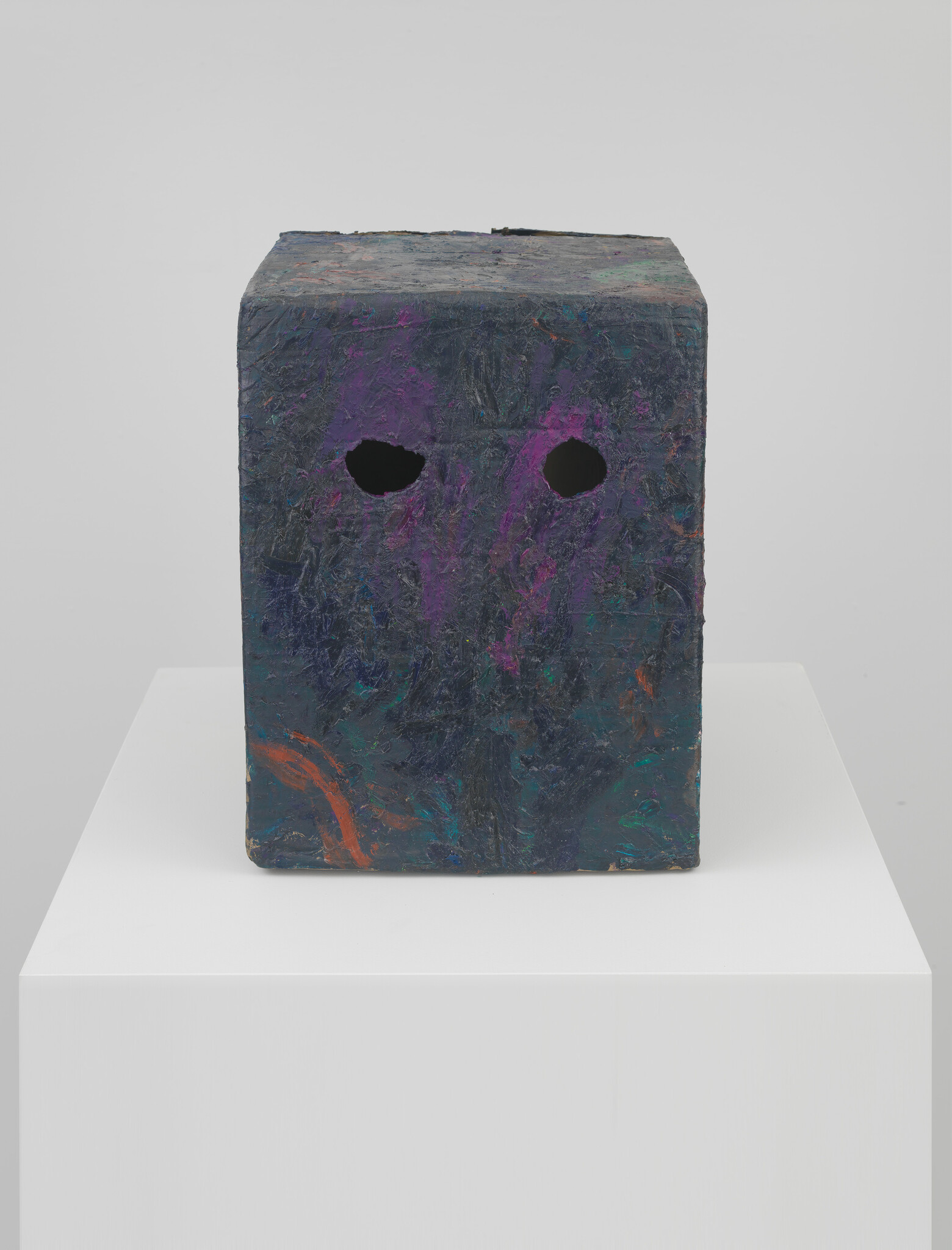 Dark painted cube sculpture with two eye-like holes sits on a white pedestal.
