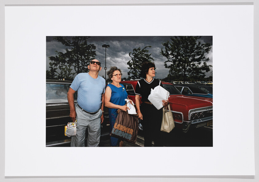 Three people stand by cars in a parking lot holding shopping bags and looking up at the sky.