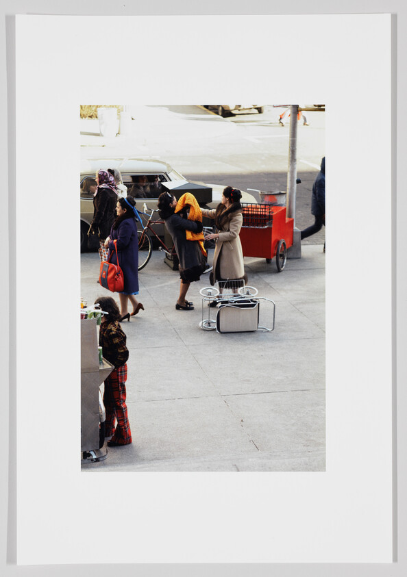 Three women stand on a sidewalk exchanging a bright yellow coat near a red street cart.