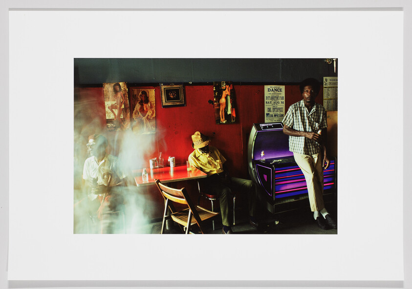 Two men sit and one leans by a jukebox in a smoky, dimly lit bar.