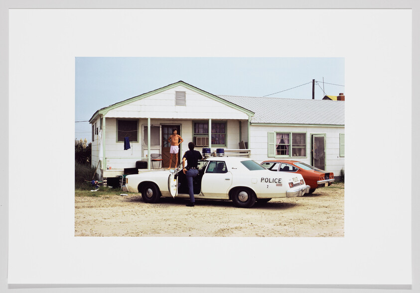 A police officer speaking to a shirtless man on a porch beside a patrol car.