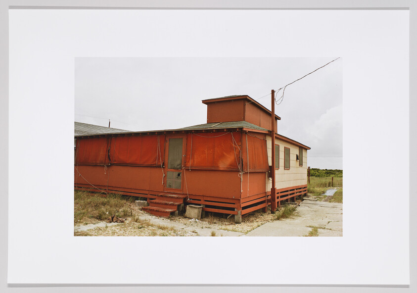 A weathered red trailer with covered porch and small steps sits on sandy ground near a utility pole.