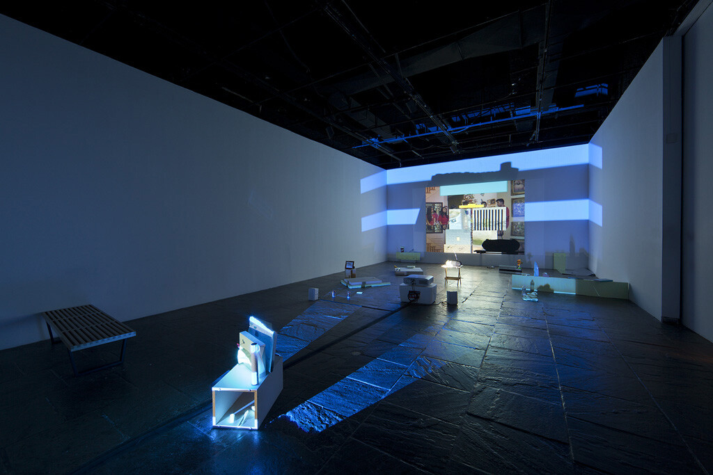 Dark gallery with scattered small sculptures and blue light projections across the floor and far wall.