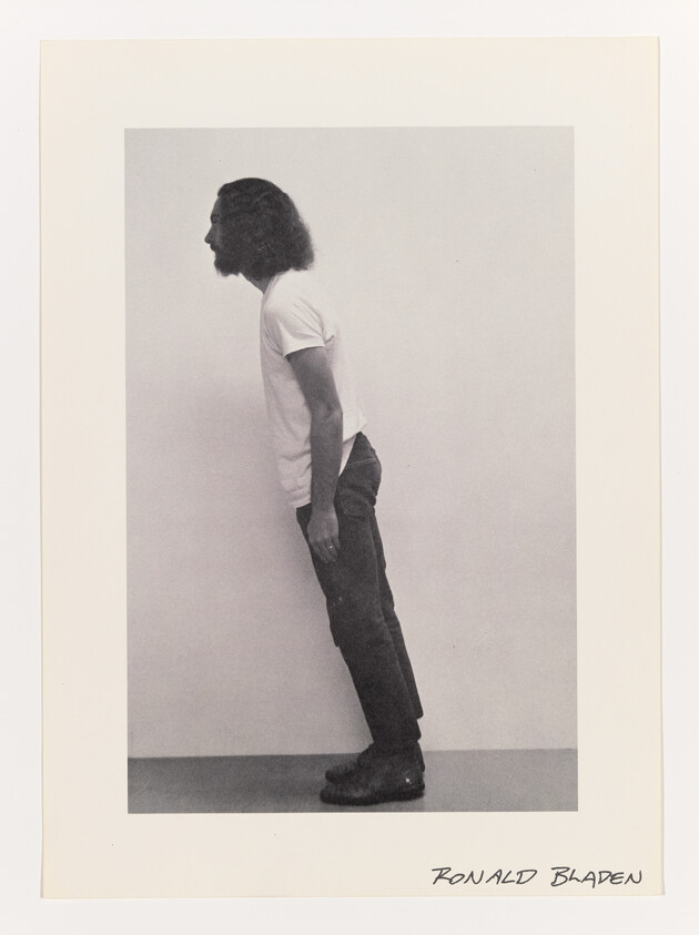 Man with long hair leans forward at an angle against a plain wall.